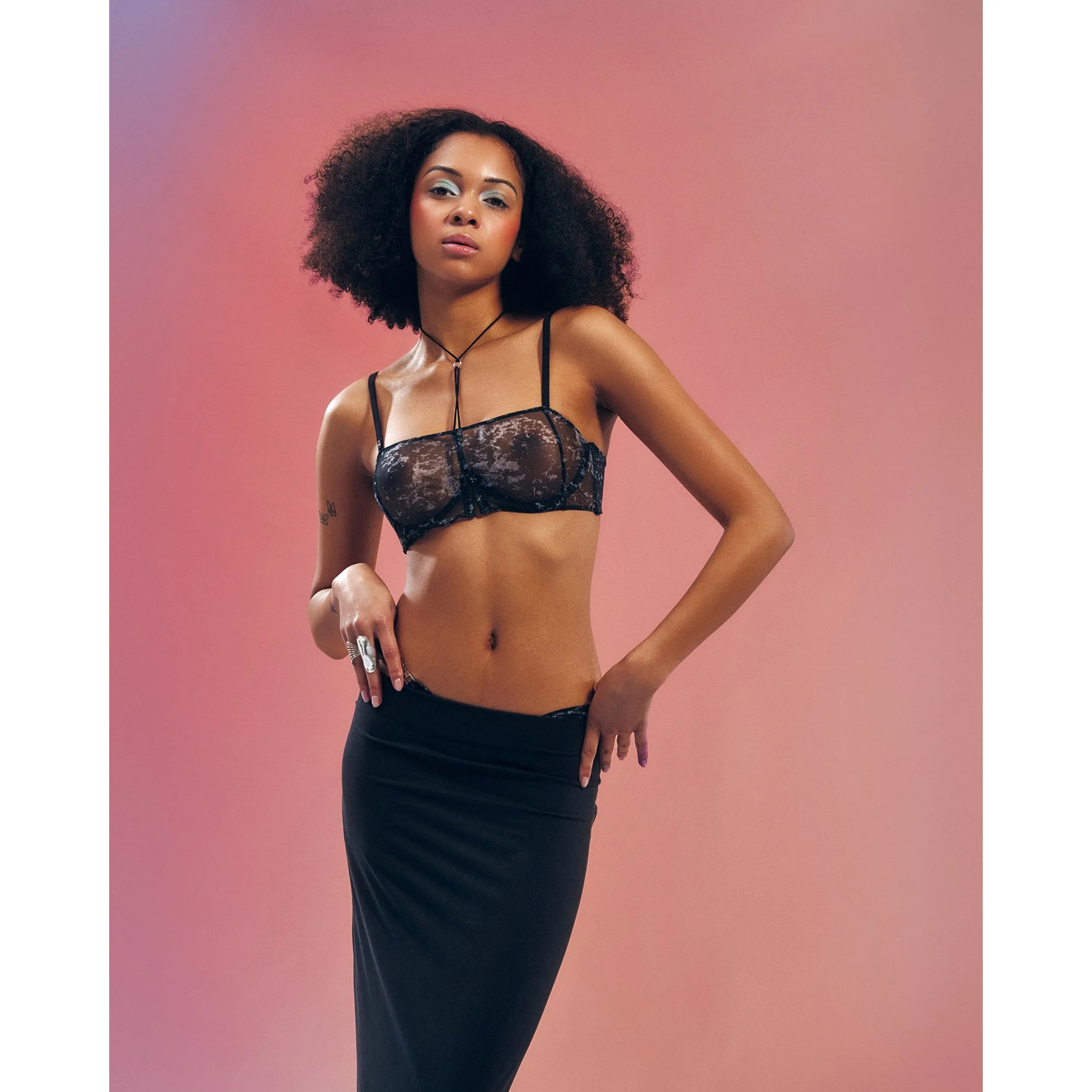 Soutien-gorge Corrosis noir transparent froncé, armatures réglables, motif raffiné, porté par mannequin sur fond rose.