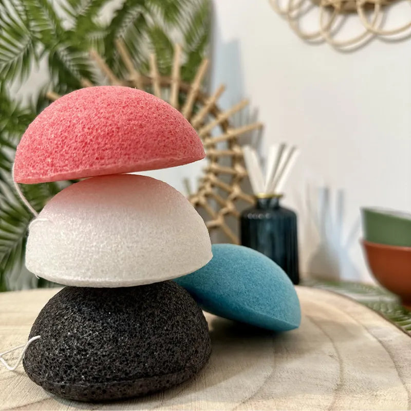Éponges konjac colorées, noire au charbon pour peau grasse, sur table en bois, fond végétal.