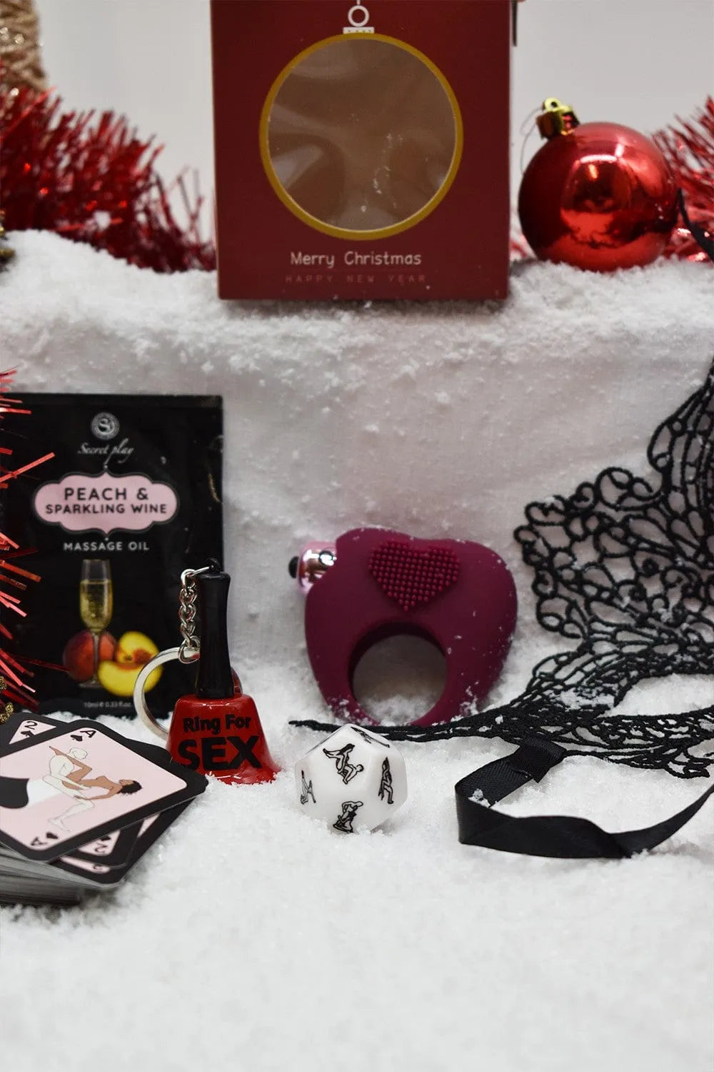 Coffret coquin Noël: huile massage pêche, cockring cœur, masque brodé, cartes, sur fond festif neige et décorations rouges.