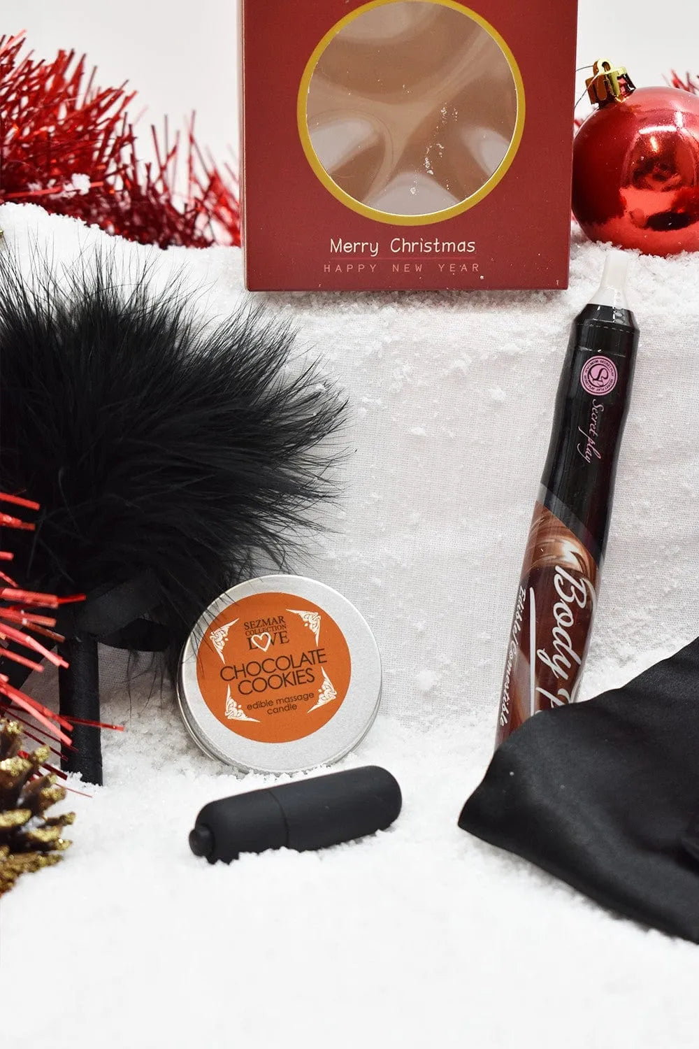 Coffret cadeau Noël chocolaté avec plumeau noir, vibromasseur, cookies chocolat, et huile comestible sur fond neigeux.