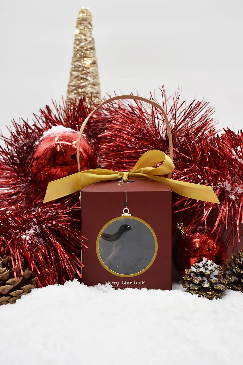 Coffret cadeau rouge avec ruban doré, entouré de décorations de Noël rouges et dorées, sur fond de neige.