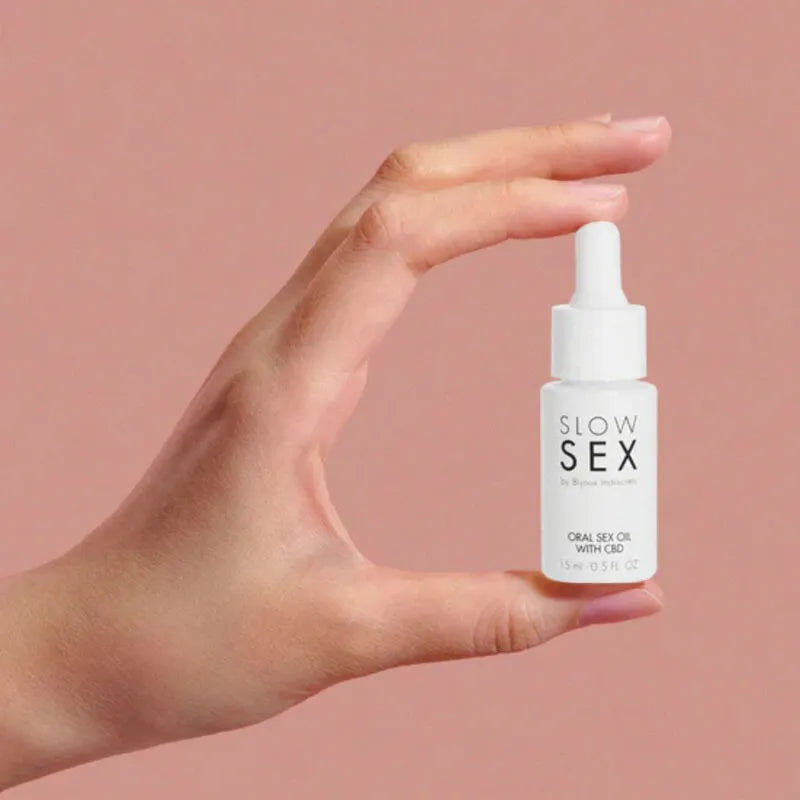 Flacon d'huile de sexe oral au CBD Slow Sex, 15 ml, blanc, avec pipette, tenu par une main sur fond rose.