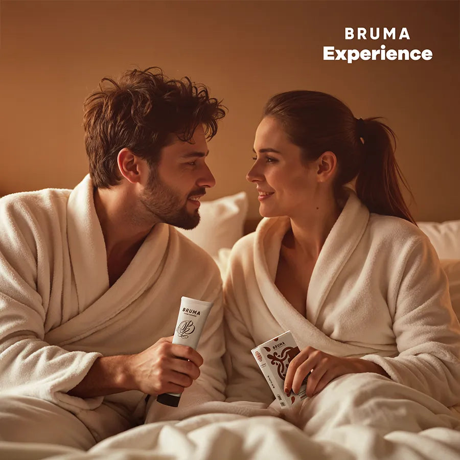 Couple en peignoirs blancs avec coffret intime BRUMA, gel lubrifiant et accessoires sur lit, ambiance chaleureuse.