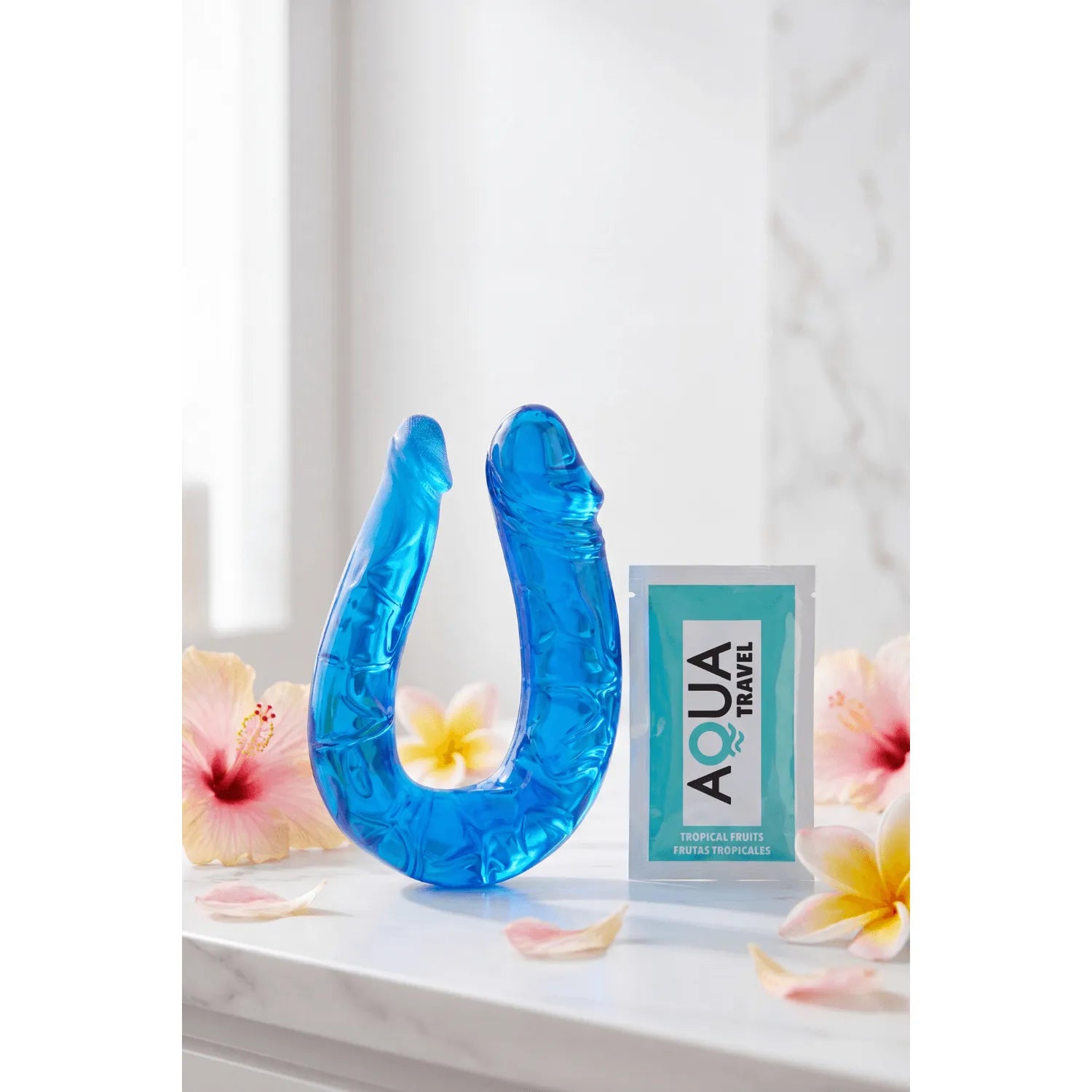 Double dong flexible bleu translucide en TPE, 26cm, stimulation réaliste, posé sur une surface avec fleurs et sachet Aqua.