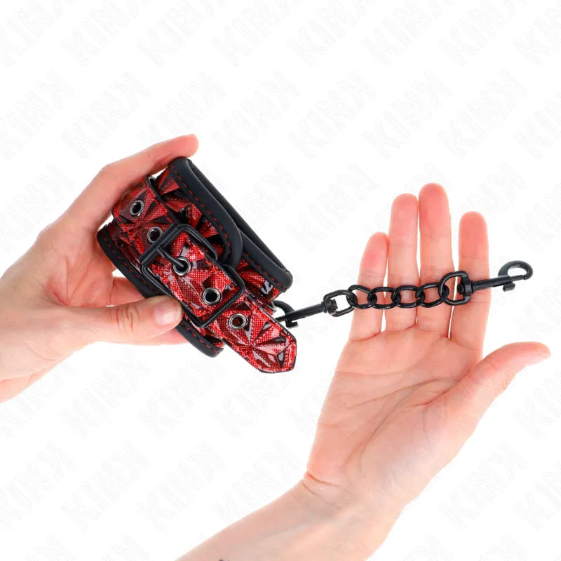Bandes de poignet BDSM rouges en cuir végétalien, ajustables, avec anneaux et chaîne noire, tenues par des mains.