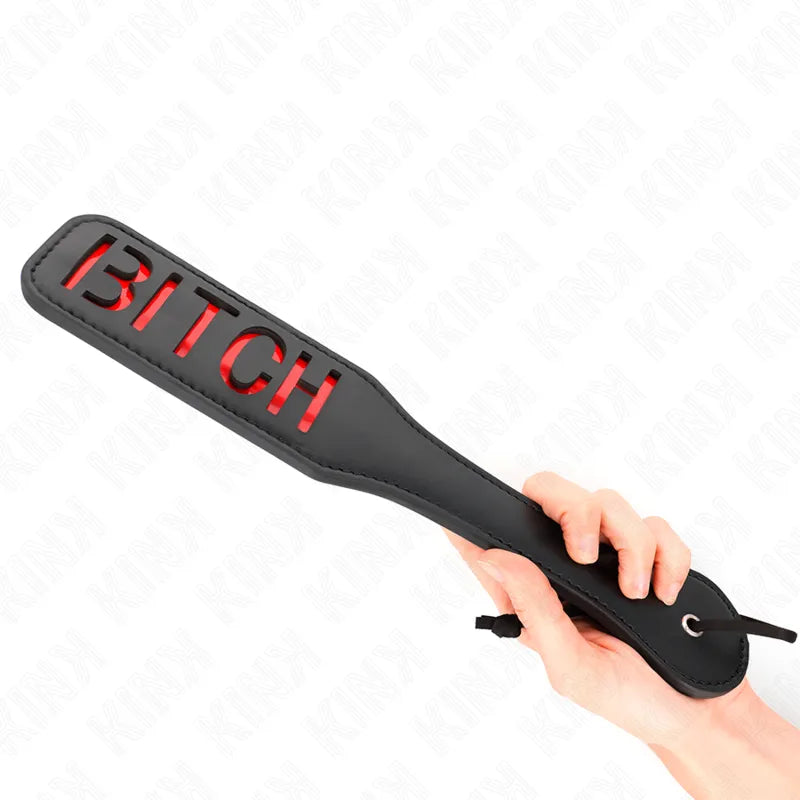 Accessoire BDSM pagaie noire en cuir avec découpe rouge BITCH, tenue par une main, pour jeux érotiques intenses.