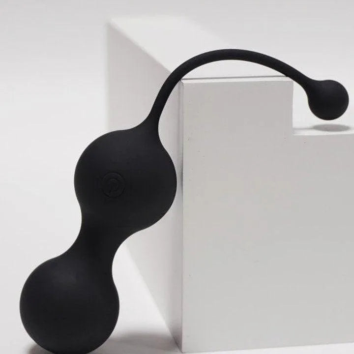 Boules de Geisha noires en silicone, forme ergonomique, avec télécommande, posées sur un fond blanc minimaliste.