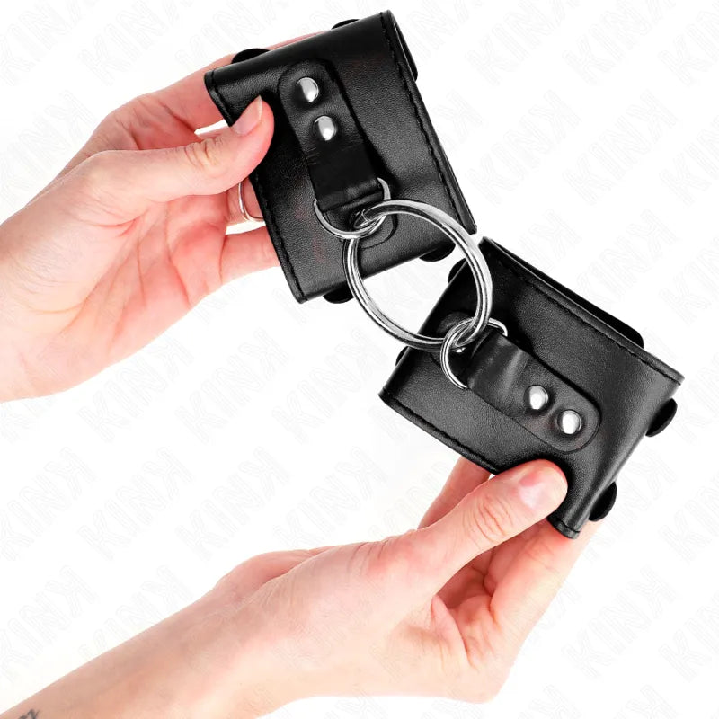 Bracelets BDSM noirs en cuir végétalien, rembourrés, avec attaches métalliques robustes, tenus par des mains.