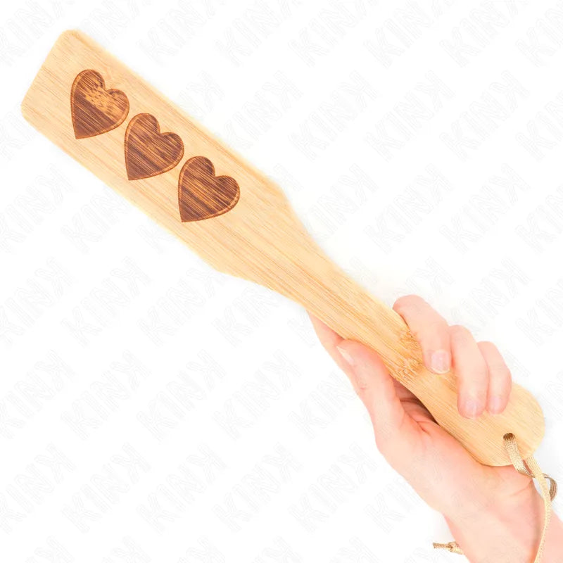 Palette en bois avec trois cœurs gravés, tenue en main, accessoire BDSM artisanal pour jeux de domination.